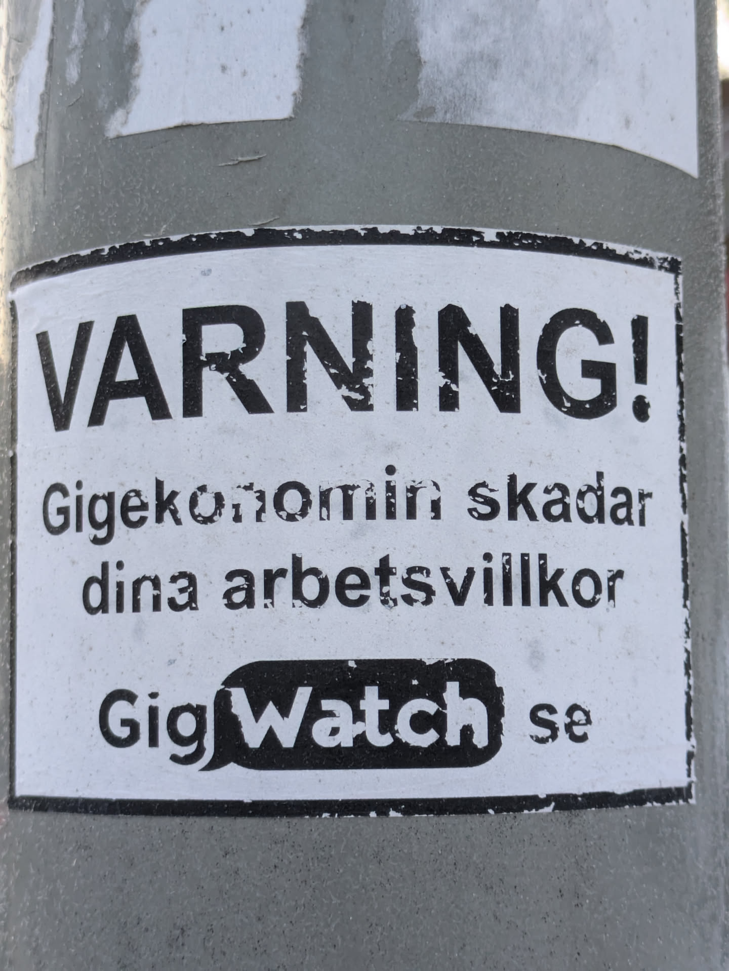 A black and white warning sticker about the gig economy's negative impact on working conditions. It features the text 'WARNING!' in large bold letters, followed by a message in Swedish and the website GigWatch.se.