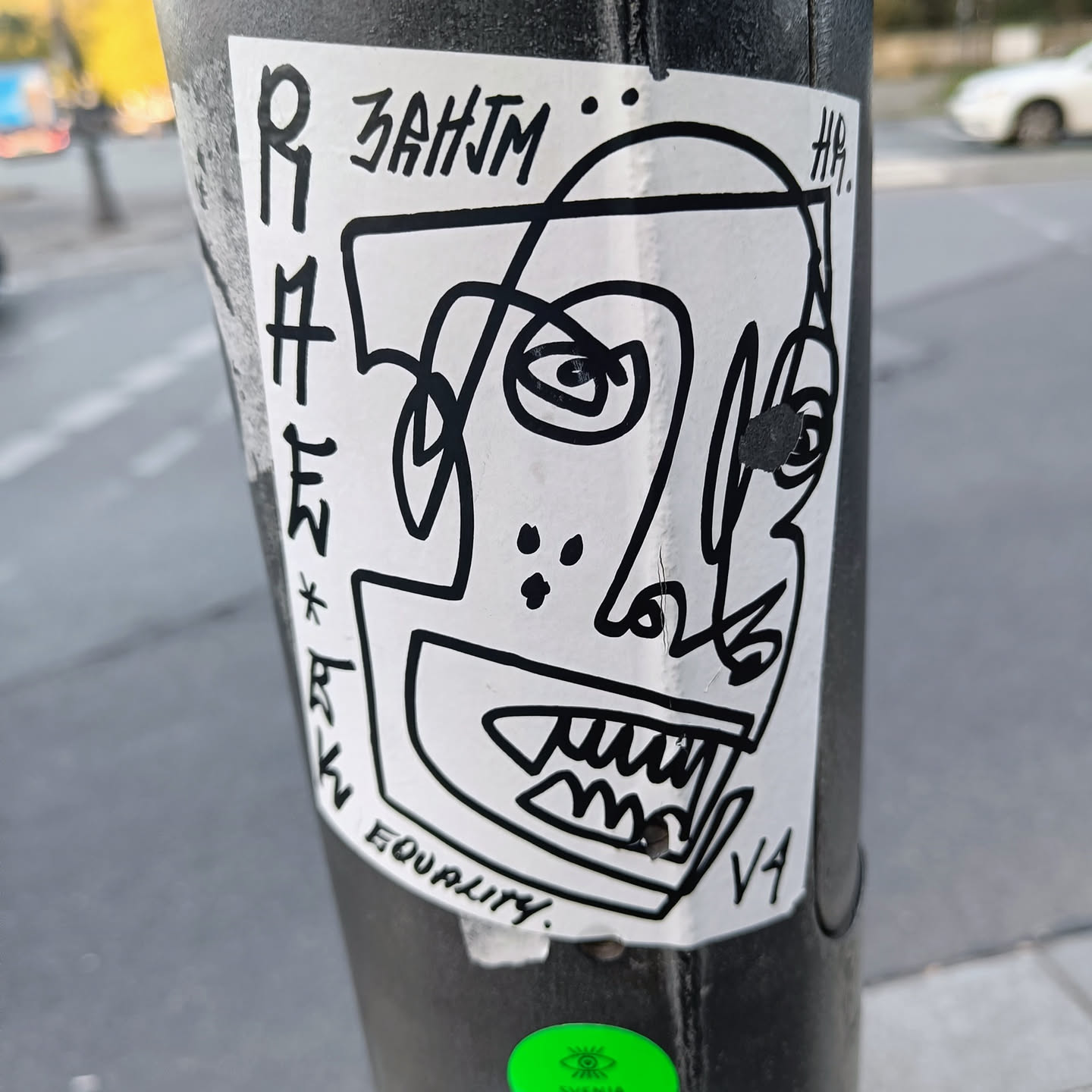 Black and white sticker featuring a stylized, abstract face with sharp lines and bold features. The style is reminiscent of cubism or graffiti art.  The sticker includes the text "ZAHJM" at the top and "EQUALITY" at the bottom. There are also some illegible markings next to the face illustration. The sticker is affixed to a pole.
