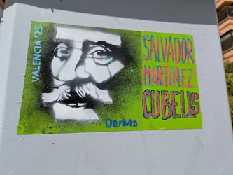 A green sticker adhered to a wall features a black and white stencil portrait of Salvador Martinez Cubells, a Spanish painter from the 19th century. The text "VALENCIA '25" is written vertically on the left side. The artist's name is written in a graffiti-style font in pink and blue, and "DarMa" is written below the portrait in blue. The sticker is bright and eye-catching.