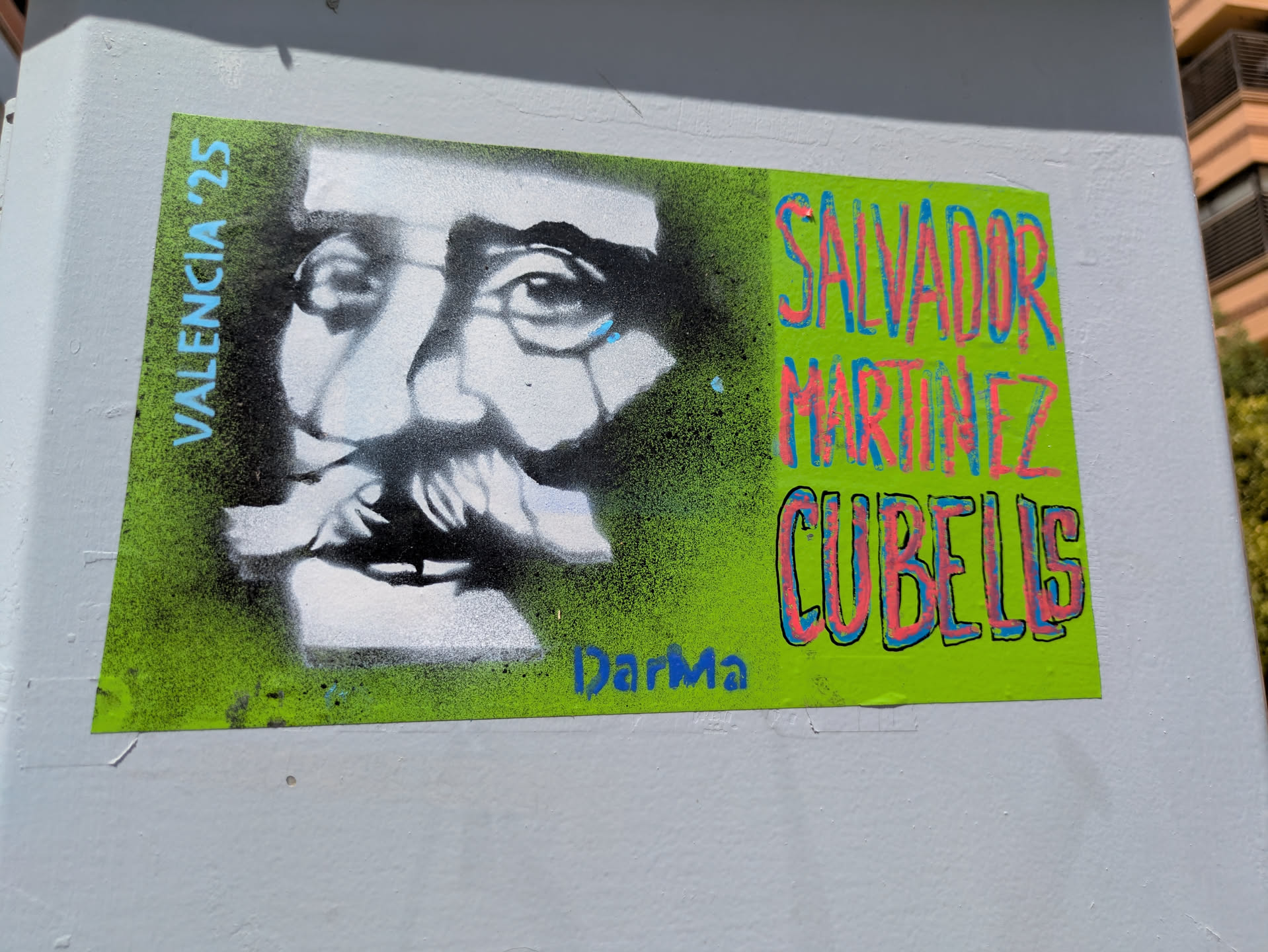 A green sticker adhered to a wall features a black and white stencil portrait of Salvador Martinez Cubells, a Spanish painter from the 19th century. The text "VALENCIA '25" is written vertically on the left side. The artist's name is written in a graffiti-style font in pink and blue, and "DarMa" is written below the portrait in blue. The sticker is bright and eye-catching.