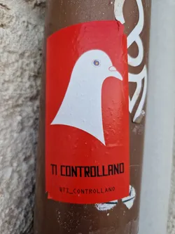 A red sticker with a white stylized dove and the text "TI CONTROLLANO" (meaning "They are controlling you") in black.  The Instagram handle @ti_controllano is also present at the bottom. The sticker is affixed to a brown pole.