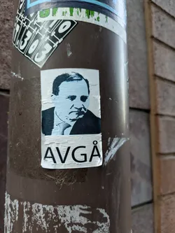 A sticker featuring a stylized black and white portrait of a man with the word "AVGÅ" written below.  The word likely means "resign" in Swedish.