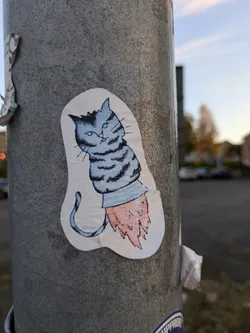 A charming sticker depicting a cartoon cat riding a rocket. The cat is blue and white with a striped tail, while the rocket is a simple, yet effective design. The artist's signature is subtly present at the bottom of the sticker. 