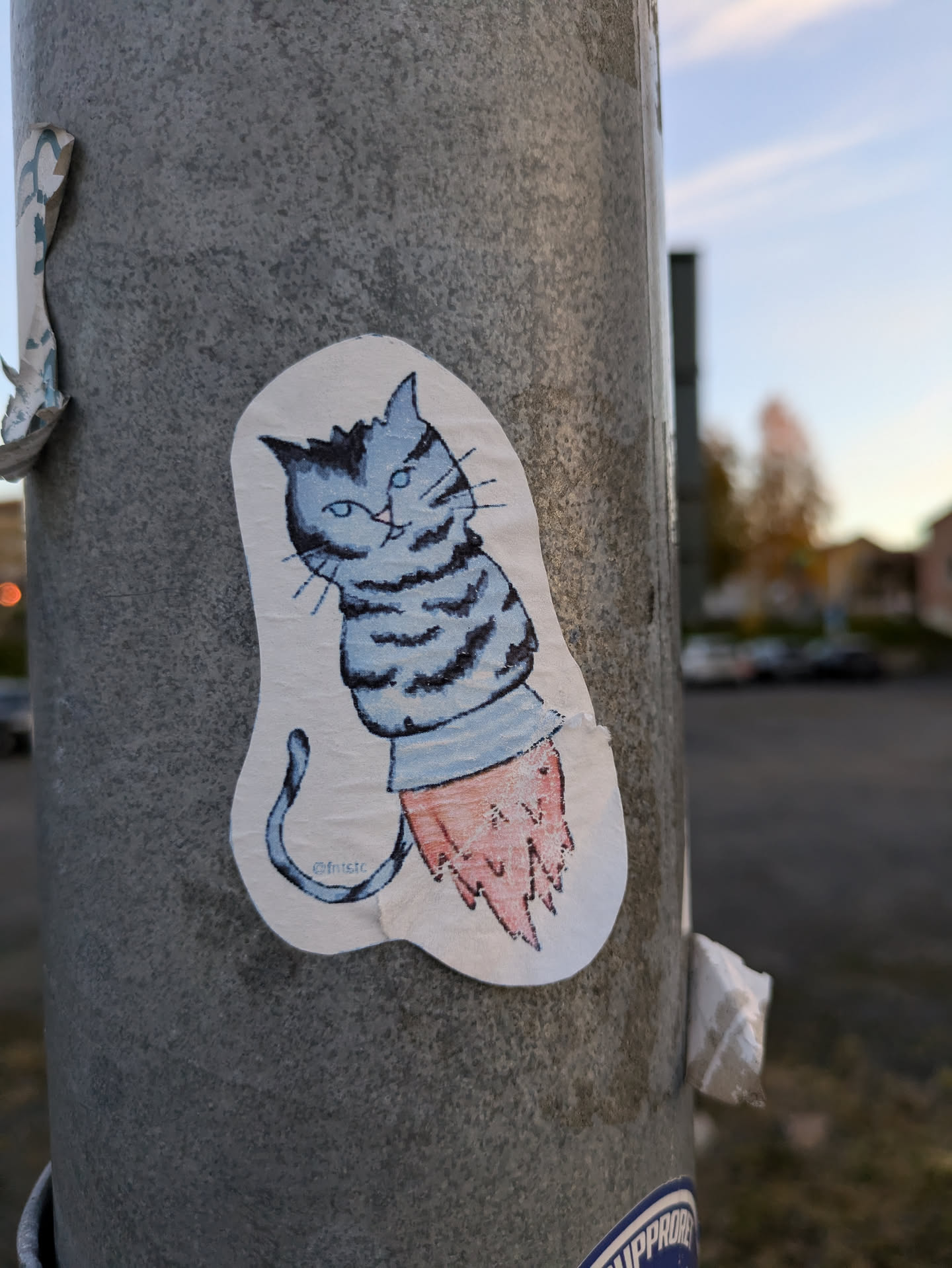 A charming sticker depicting a cartoon cat riding a rocket. The cat is blue and white with a striped tail, while the rocket is a simple, yet effective design. The artist's signature is subtly present at the bottom of the sticker. 