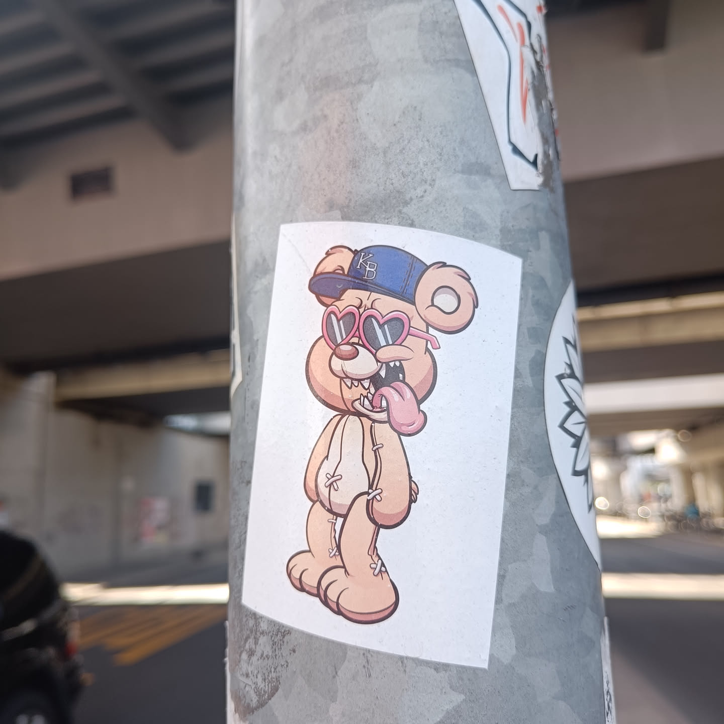 Sticker of a cartoon bear wearing heart-shaped sunglasses and a baseball cap. The bear has its tongue sticking out. 