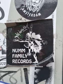 A black and white sticker featuring the logo for Numm Family Records. The logo includes a stylized figure in a dancing pose. The sticker is slightly damaged around the edges.