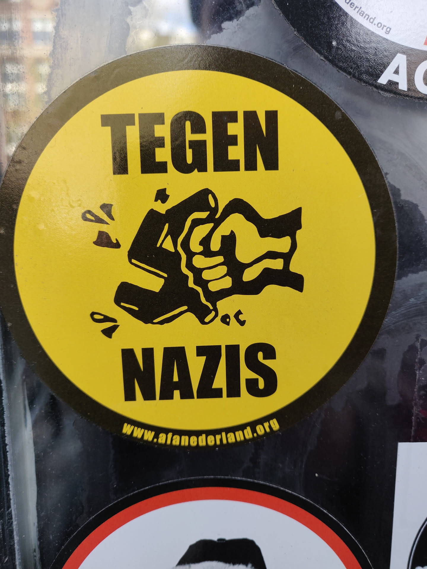 A round yellow sticker with a black fist smashing a swastika. The text 'TEGEN NAZIS' is written above and below the image.  The website www.afaneederland.org is also visible at the bottom.