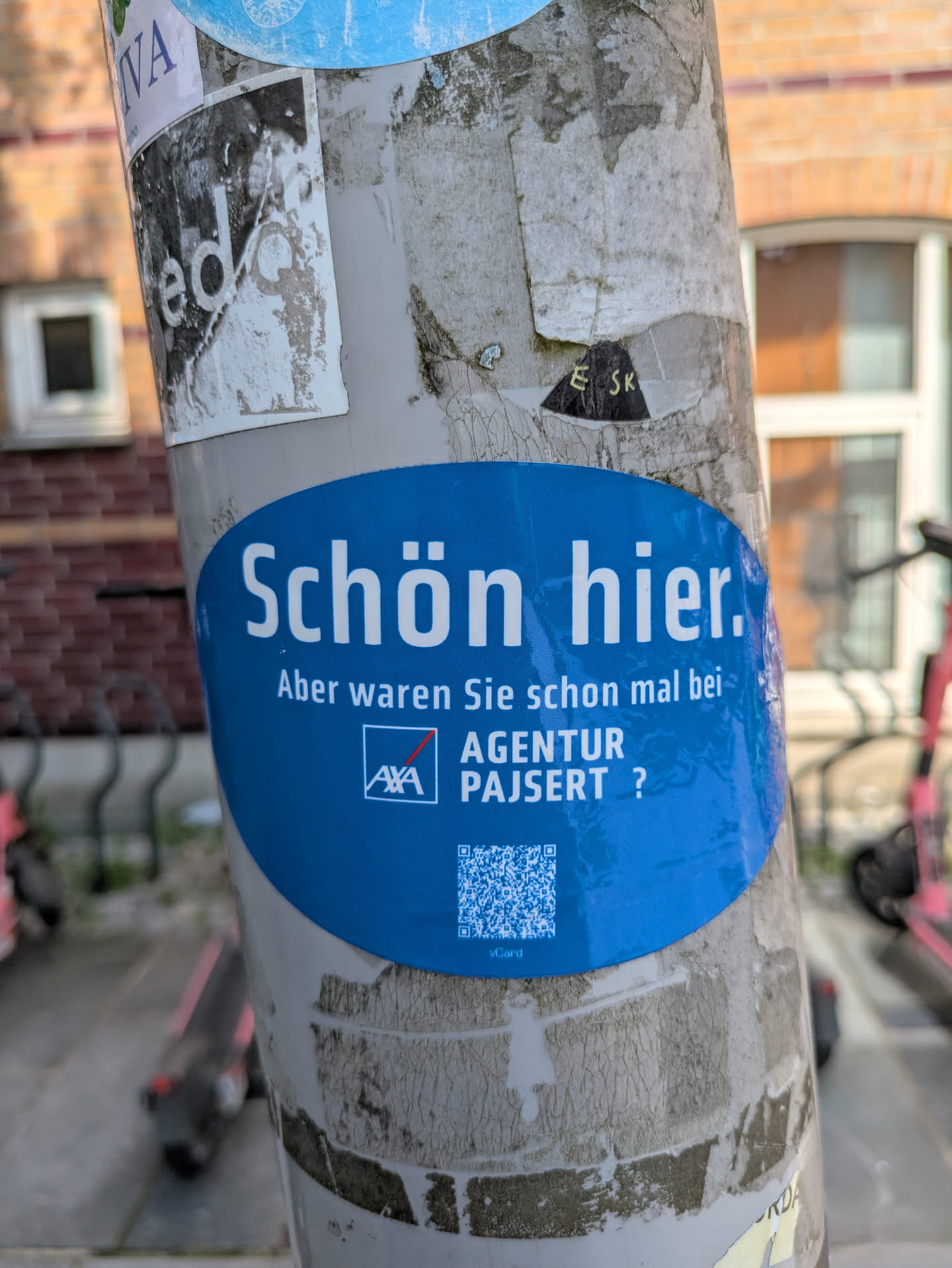 A blue oval-shaped sticker is affixed to a weathered utility pole, which is also covered with remnants of other stickers. The sticker's design is clean and features white text on a solid blue background. The main text, in German, reads "Schön hier. Aber waren Sie schon mal bei- AXA AGENTUR PAJSERT ?" which translates to "It's nice here. But have you ever been to- AXA AGENCY PAJSERT ?". Below the agency name, there is a clear QR code with the small text "vCard" underneath it, indicating it's for contact information. The pole itself shows signs of age and previous adhesive marks, typical of urban environments.