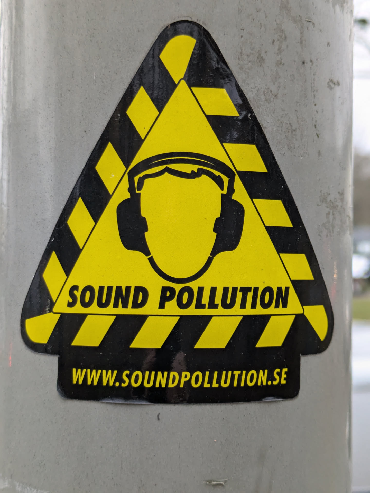 A triangular yellow and black sticker with a person wearing earmuffs in the center and the text 'SOUND POLLUTION' and 'WWW.SOUNDPOLLUTION.SE'.