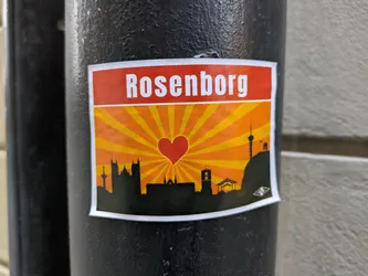 A sticker depicting a stylized skyline of Rosenborg, Denmark, with a prominent heart at its center, set against a radiant orange sunburst background. The sticker features a minimalist design with the city's iconic buildings and landmarks in silhouette.