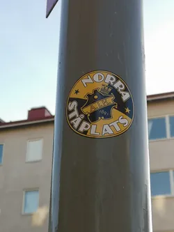 A circular sticker predominantly in yellow and black, featuring a central crest. The crest is black with yellow details, including a stylized sun, a crown, and the letters 'A.I.K.' with the year '1891' beneath them. Surrounding the crest, along the top curve, is the word 'NORRA' in white capital letters with a black outline, set against a yellow background. Along the bottom curve, also in white capital letters with a black outline on a yellow background, is 'STÅPLATS'. Two yellow stars are positioned on the yellow background, one on each side, separating the 'NORRA' and 'STÅPLATS' text.