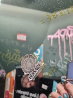 A small, round Versace sticker featuring the iconic Medusa head logo. The sticker is affixed to a glass surface with graffiti in the background.  The word VERSACE is also printed on a separate piece of the sticker.