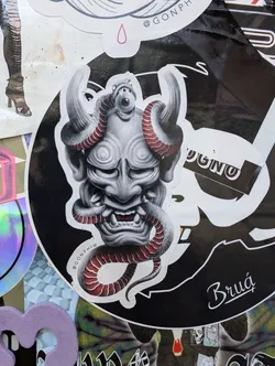 Round sticker featuring a detailed illustration of an oni mask entwined with a red snake. The style is dark and graphic with shades of grey and red. The artist's Instagram handle is visible in small text at the bottom. 