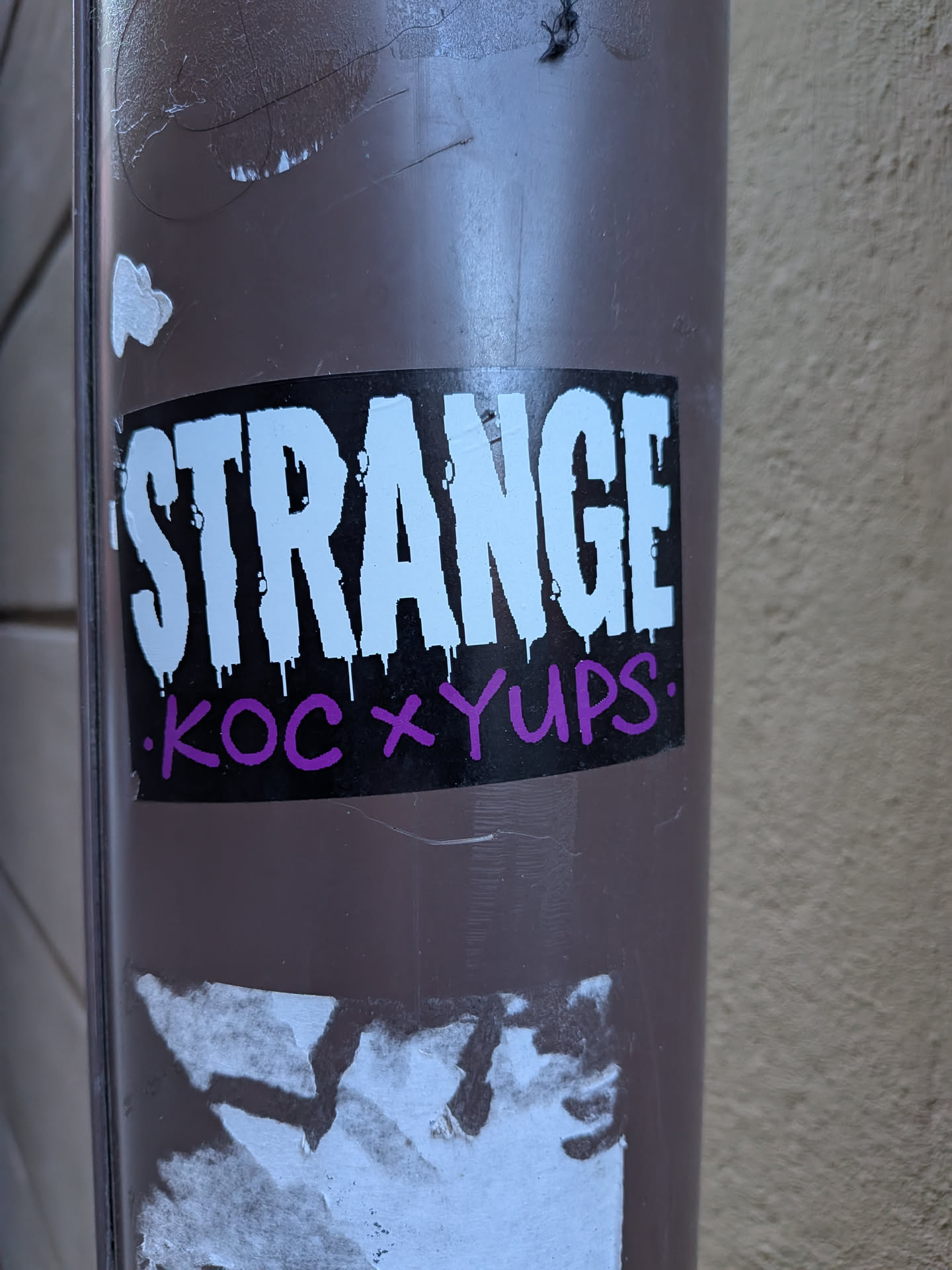 A rectangular sticker with a solid black background. The word "STRANGE" is prominently displayed in large, bold, white capital letters, featuring a dripping paint effect at the bottom of each character. Below this, in smaller, purple, handwritten-style capital letters, is the text "KOC x YUPS.", flanked by small white dots. The sticker is applied to a brown pole, showing some wear and tear, with remnants of other stickers visible below it.