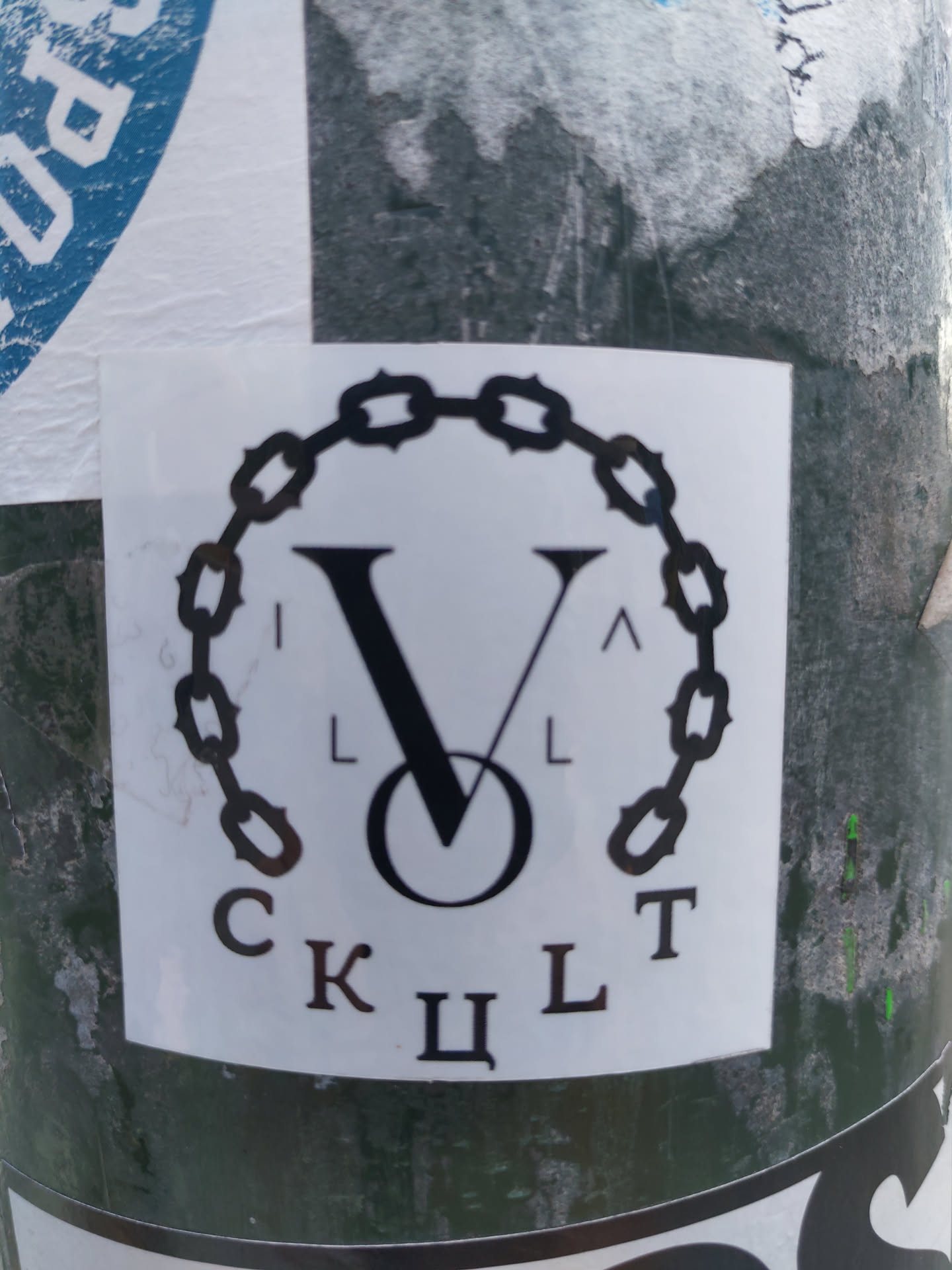 A black and white sticker with a logo featuring a large V enclosed within a circular chain. The Cyrillic letters 'CKЦЛT' are positioned below the logo.  The sticker is affixed to a dark green surface, possibly a pole or pillar. 