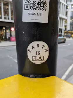 A circular, off-white or cream-colored sticker adhered to a textured black pole. The sticker features dark brown or black text in a classic serif font. The word 'EARTH' is curved along the top inner edge of the circle, 'IS' is centered below it, and 'FLAT' is centered at the bottom, framed by two horizontal lines. The sticker shows minor signs of wear, consistent with outdoor placement.