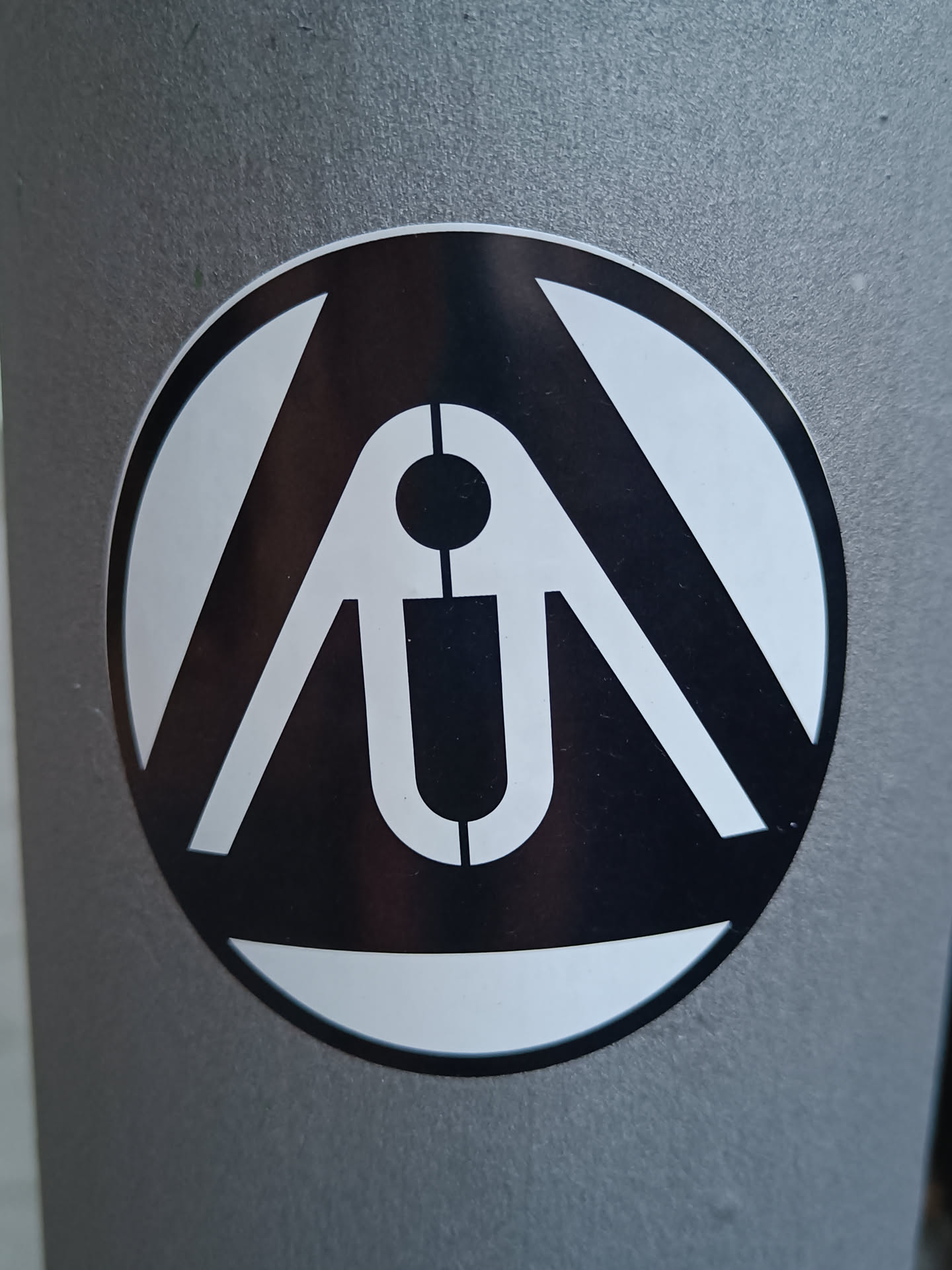 A round sticker with a black and white design. The logo appears to be an abstract symbol or emblem. The sticker is affixed to a gray surface, possibly a pole or a wall.