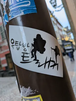 A white sticker on a brown pole with black silhouettes of two hikers walking uphill.  Korean text is present on the sticker. The style is simple and minimalistic.