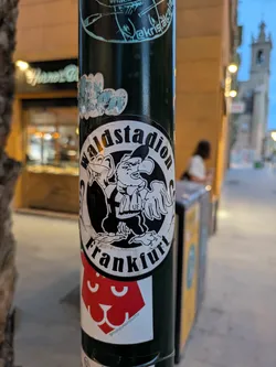 This is a black and white round sticker on a pole. The text "Waldstadion Frankfurt" wraps around a cartoon drawing with a monkey and eagle character. Below this sticker is a square one with a red abstract cat face.