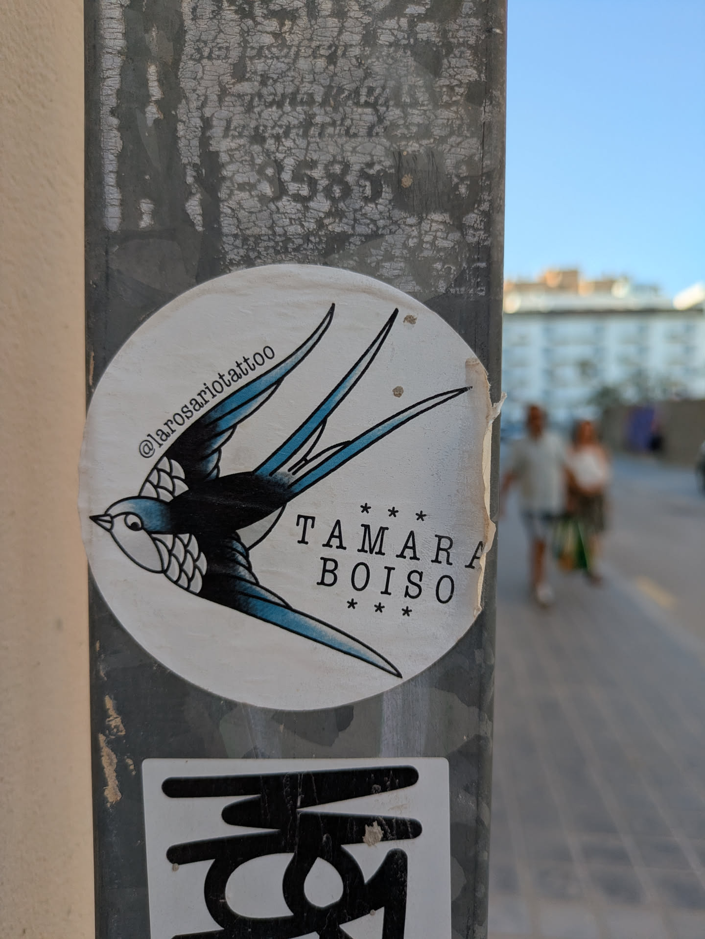 This round sticker, affixed to a metal pole, showcases a classic tattoo-style swallow. The bird is rendered in black and blue, with detailed shading on its wings and chest. The text "TAMARA BOISO" is printed in a simple font, surrounded by small stars. Above the bird, the Instagram handle @larosariotattoo is visible, indicating the artist or tattoo parlor responsible for the design.