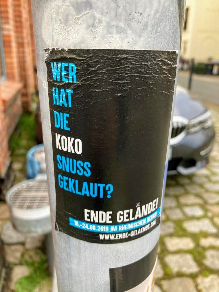 A worn-out black sticker with light blue text on a gray pole. The text is in German and asks who stole the Koko Snuss. It also mentions Ende Gelände, an event that took place from August 18th to 24th, 2019, in Rheinisches Industriemuseum.