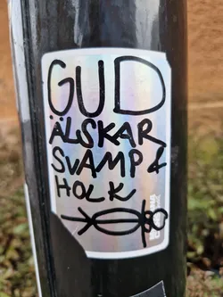 A sticker with a iridescent background and black text. The text says "Gud älskar svamp & holk" which translates from Swedish to English as "God loves mushroom & hollow". There is also a small drawing of a person under the text.