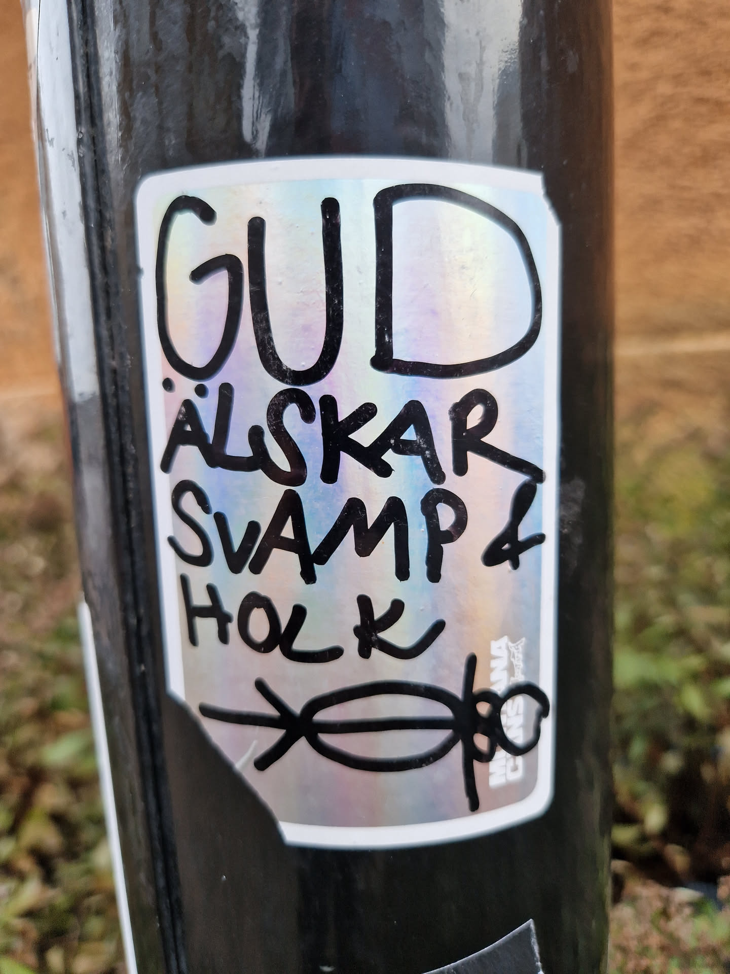 A sticker with a iridescent background and black text. The text says "Gud älskar svamp & holk" which translates from Swedish to English as "God loves mushroom & hollow". There is also a small drawing of a person under the text.
