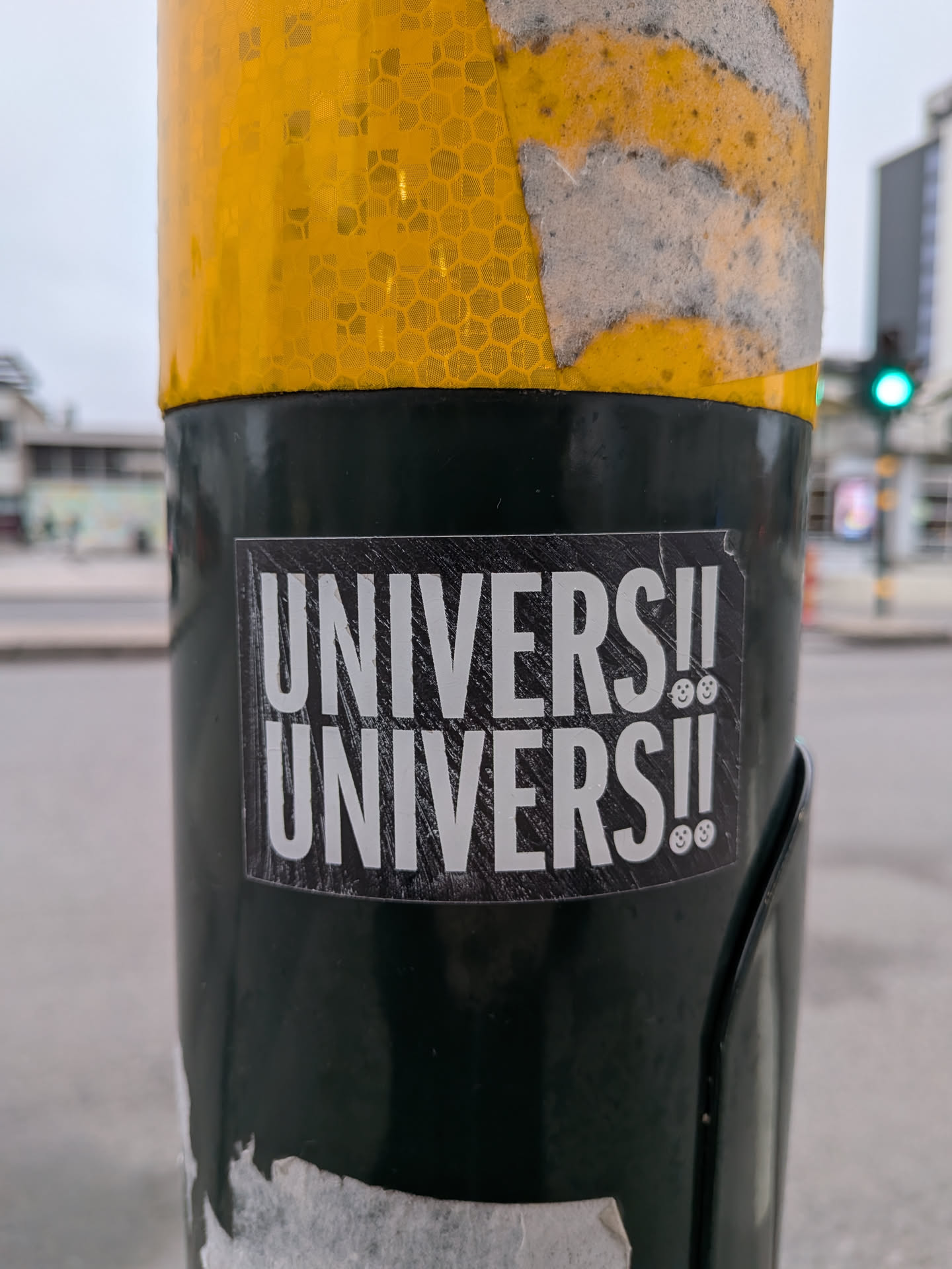 The sticker is rectangular with a black background and white text. The text reads 'UNIVERSE!!' on the top line and 'UNIVERSE!!' on the bottom line. Both lines are followed by two small smiley faces. The text has a distressed, slightly rough appearance, and the background has a subtle textured or scratched effect. The sticker is applied to a dark green cylindrical object, likely a pole.