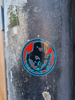 A round sticker with a black and teal design on a red background. The design appears to be an abstract figure or symbol within a circular frame of leaves or branches. There is a small heart shape near the top of the figure.