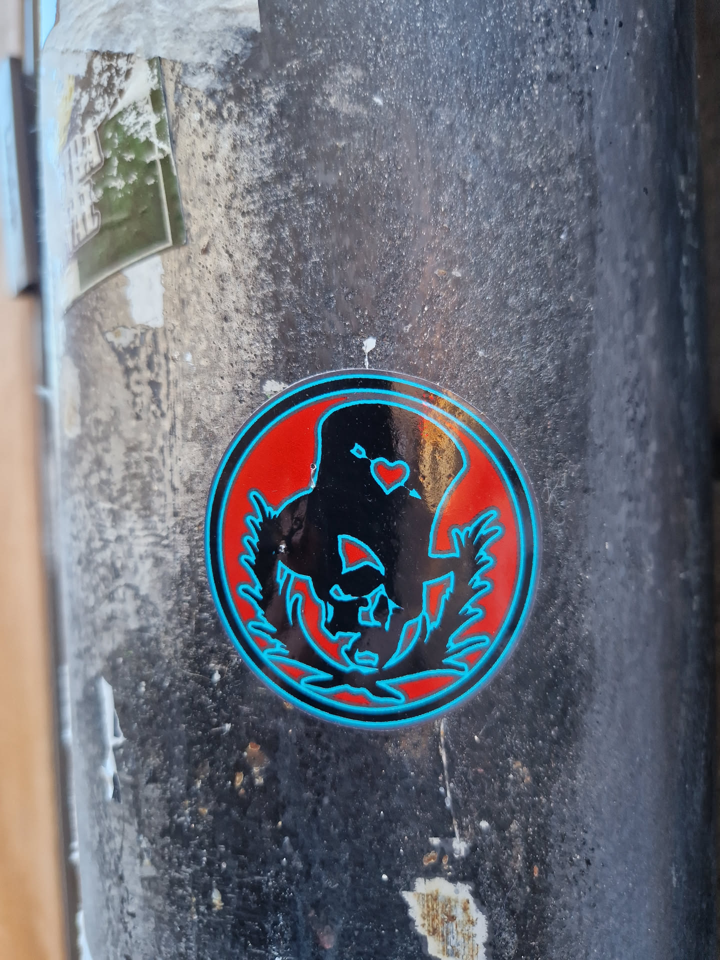 A round sticker with a black and teal design on a red background. The design appears to be an abstract figure or symbol within a circular frame of leaves or branches. There is a small heart shape near the top of the figure.