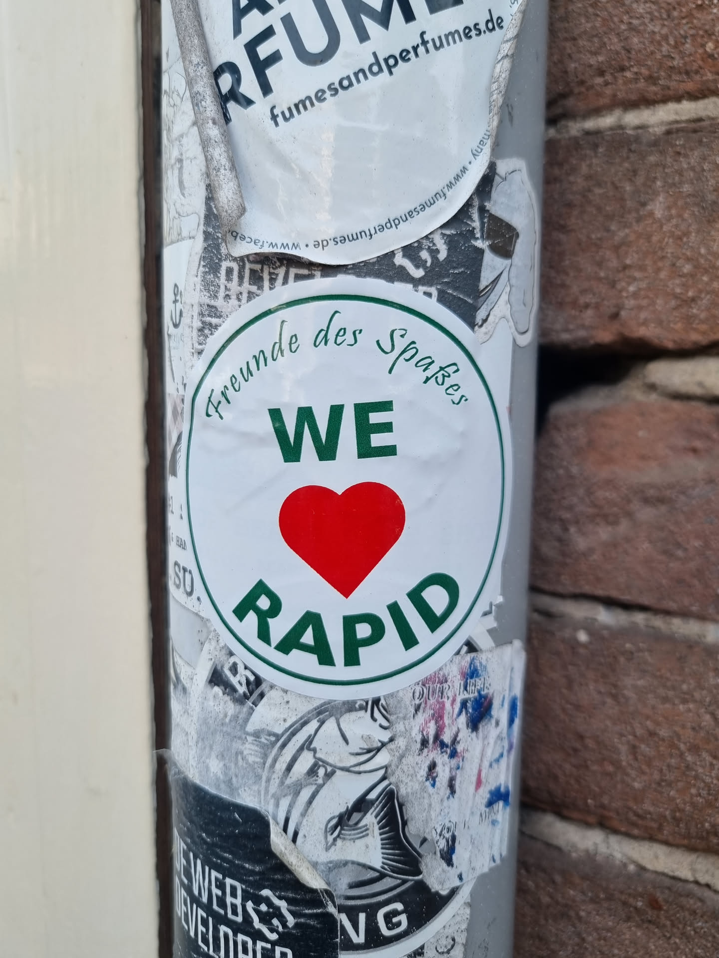 A round sticker with a green border and a red heart in the center. The text "Freunde des Spaβes" is written in a cursive font above "WE", and "RAPID" is written in a larger font below the heart. 
