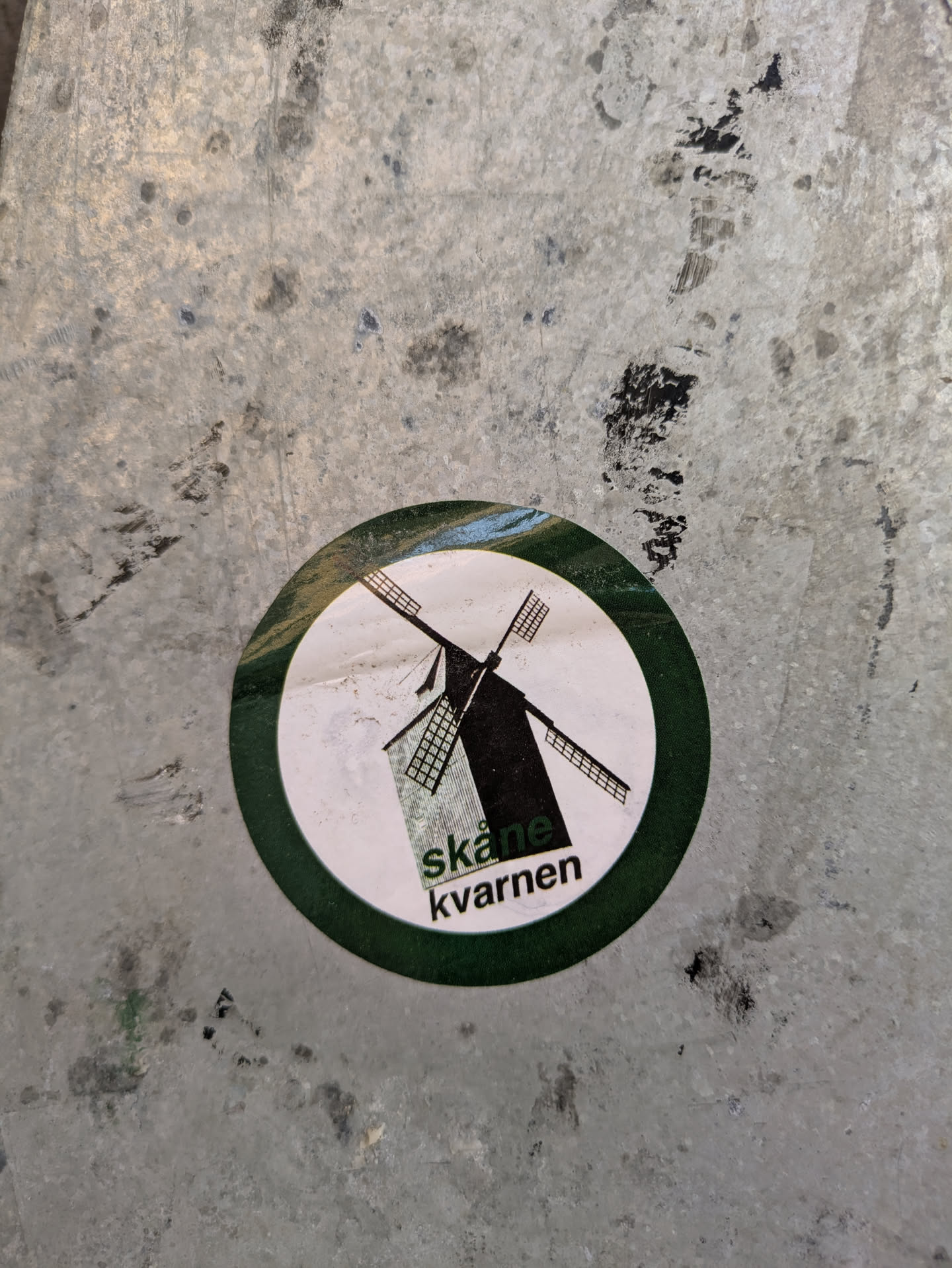 This round sticker has a white background with a green border. A black windmill graphic is in the center, and below it, the words "Skåne kvarnen" are printed in black.