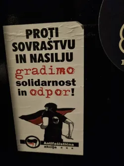 A sticker with a black and red design showing a person holding a flag. The text on the sticker is in Slovene and translates to "Against oppression and violence we build solidarity and resistance!"  It includes the logo of Antifasistična akcija.