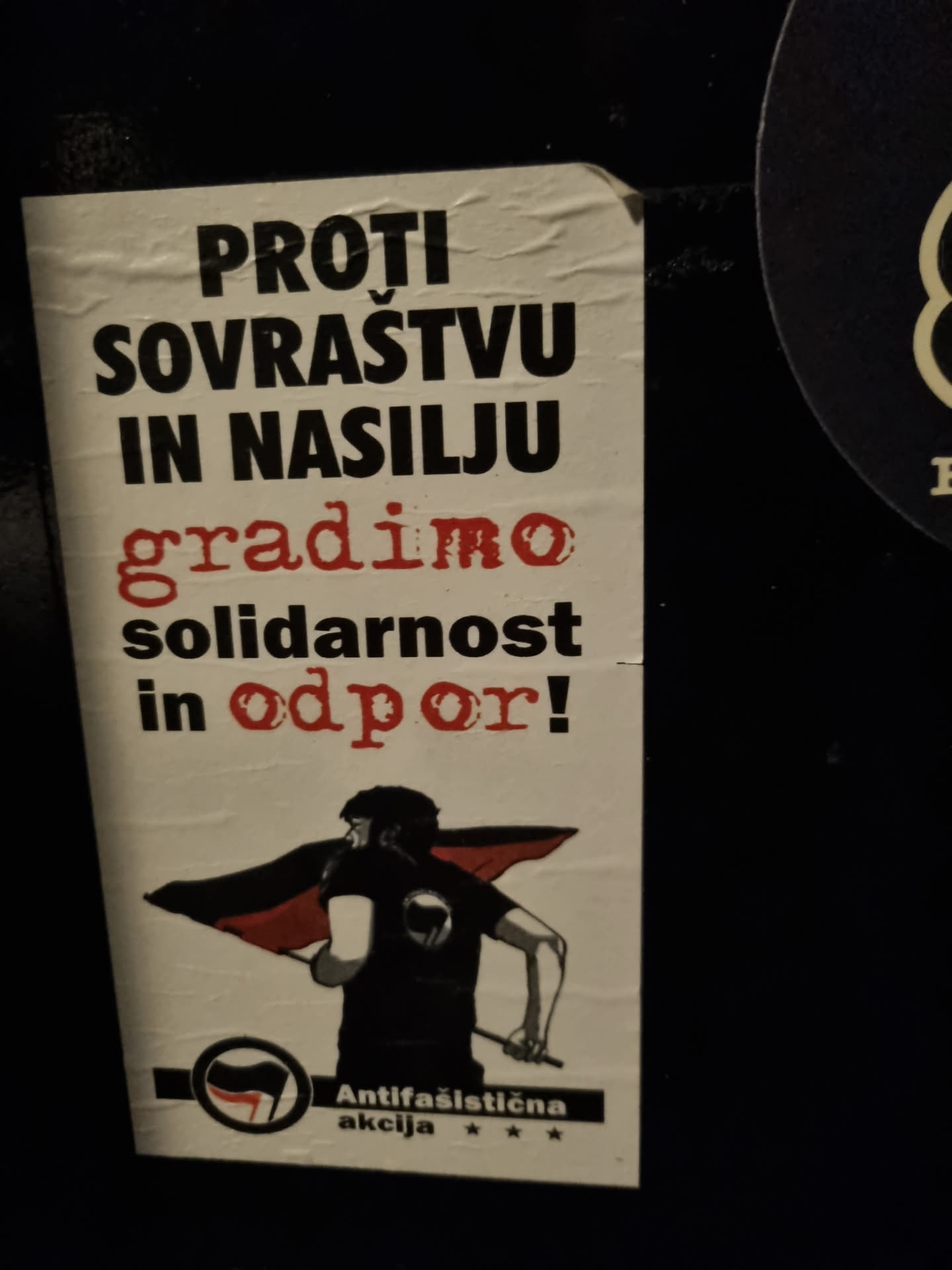 A sticker with a black and red design showing a person holding a flag. The text on the sticker is in Slovene and translates to "Against oppression and violence we build solidarity and resistance!"  It includes the logo of Antifasistična akcija.