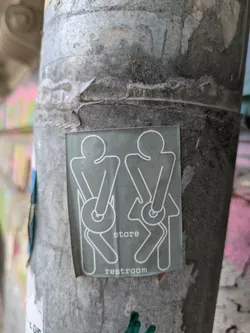 A humorous sticker on a weathered pole features stick figures holding toilet paper rolls over their rear ends. The text reads 'store restroom'.