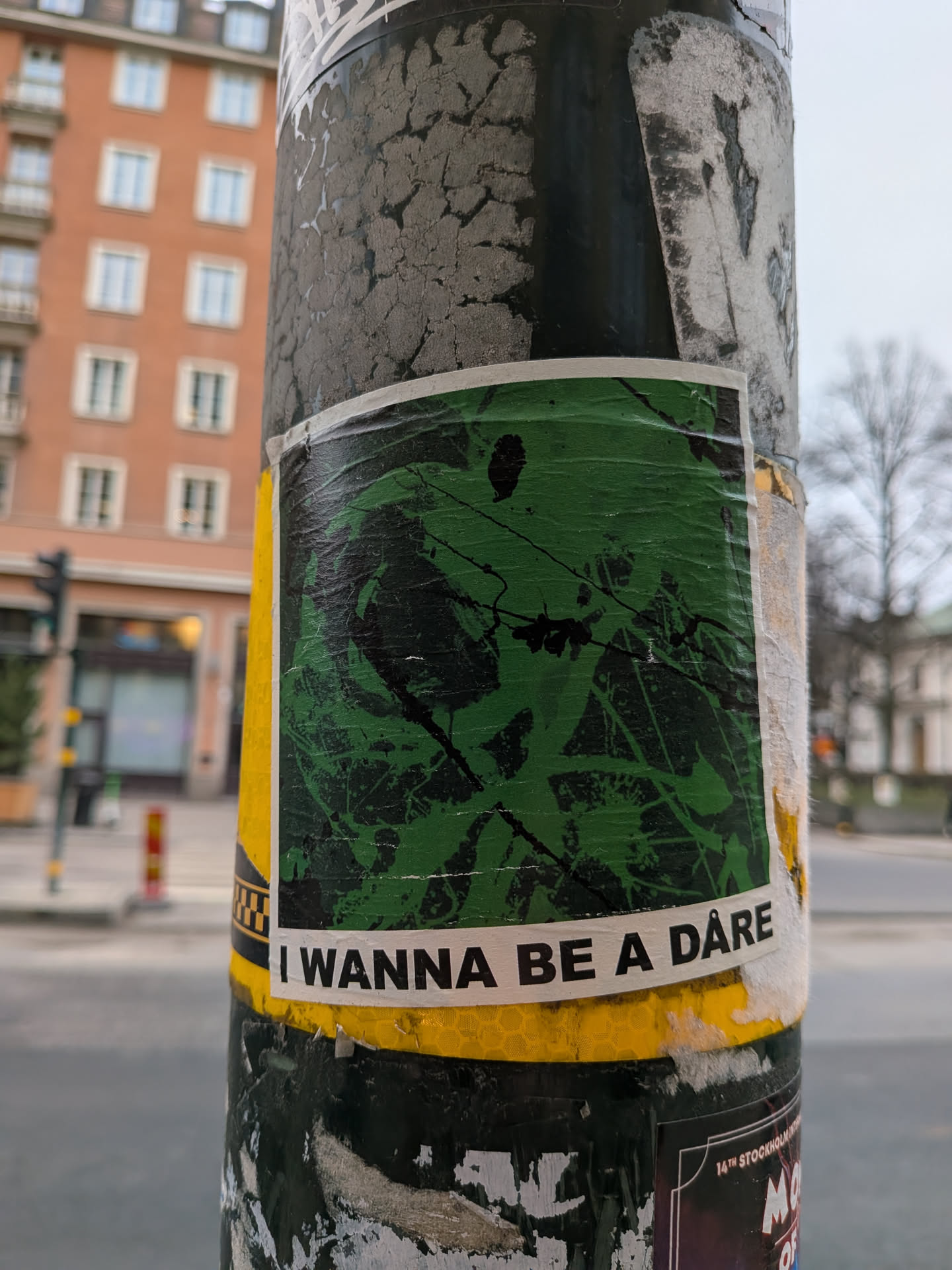 The sticker is predominantly green with abstract black splatters and lines, giving it an energetic and somewhat chaotic appearance. It has a white border and is affixed to a pole, partially covered by other worn stickers. Below the main green design, there is a yellow band with a honeycomb pattern, and a white strip with black text. The sticker appears slightly creased and weathered, suggesting it has been exposed to the elements.