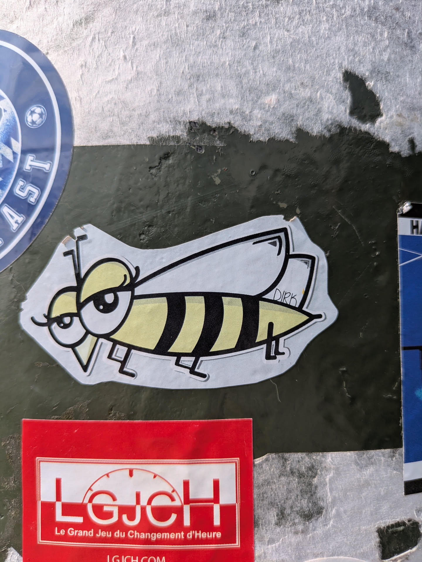 A charming cartoon sticker of a bee or wasp, featuring a yellow and black striped body, large, expressive eyes with heavy eyelids giving it a 'sassy' or 'bored' look, and delicate, transparent wings. The artist's name 'DIRK' is subtly written on one of its wings. The sticker is affixed to a textured, dark green surface, with fragments of other stickers visible around it.