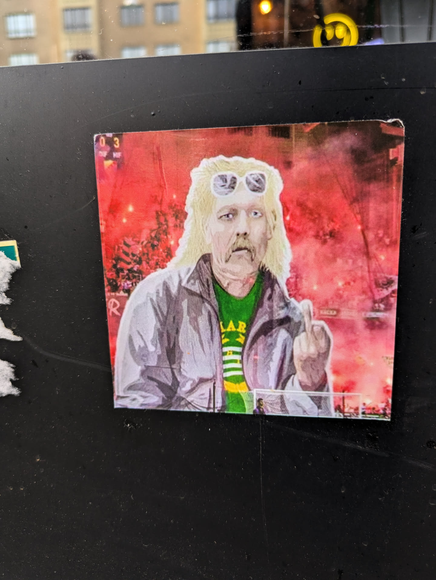 This sticker features a stylized illustration of a man who seems to be a football fan, possibly at a match where flares are being lit.  He's sporting a mullet, sunglasses, and a green and yellow shirt, giving a middle finger gesture. The style is bold and slightly graphic. The background is a hazy red, possibly suggesting a nighttime setting or the smoky atmosphere of a stadium. 