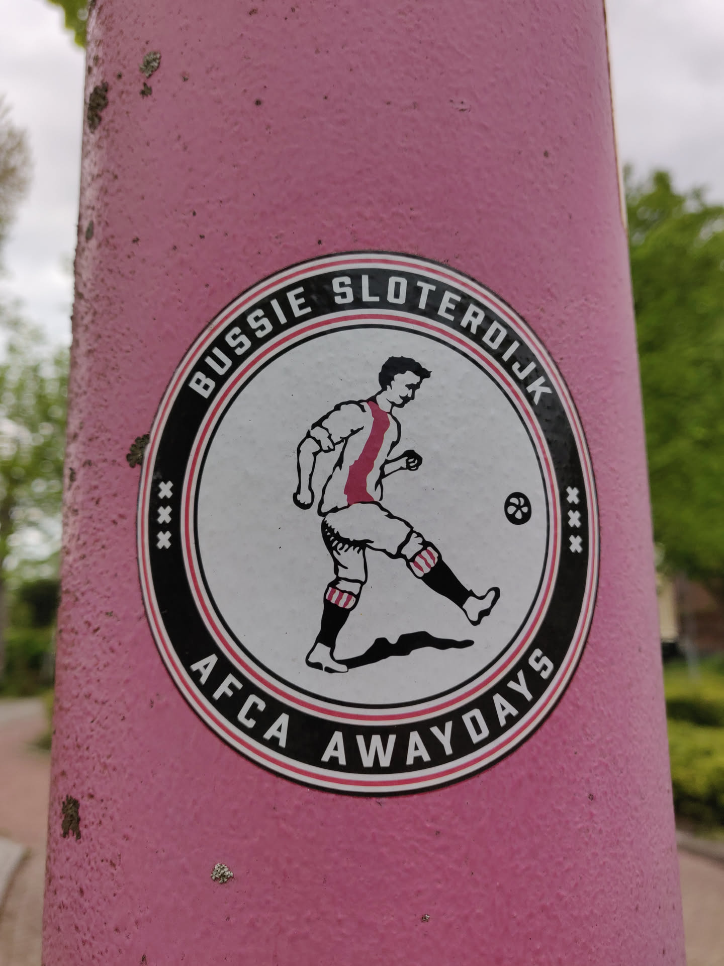 A round sticker featuring a football player in vintage attire. The text around the image says 'Bussie Sloterdijk' on the top and 'AFCA Awaydays' on the bottom. The sticker is affixed to a pink pole.
