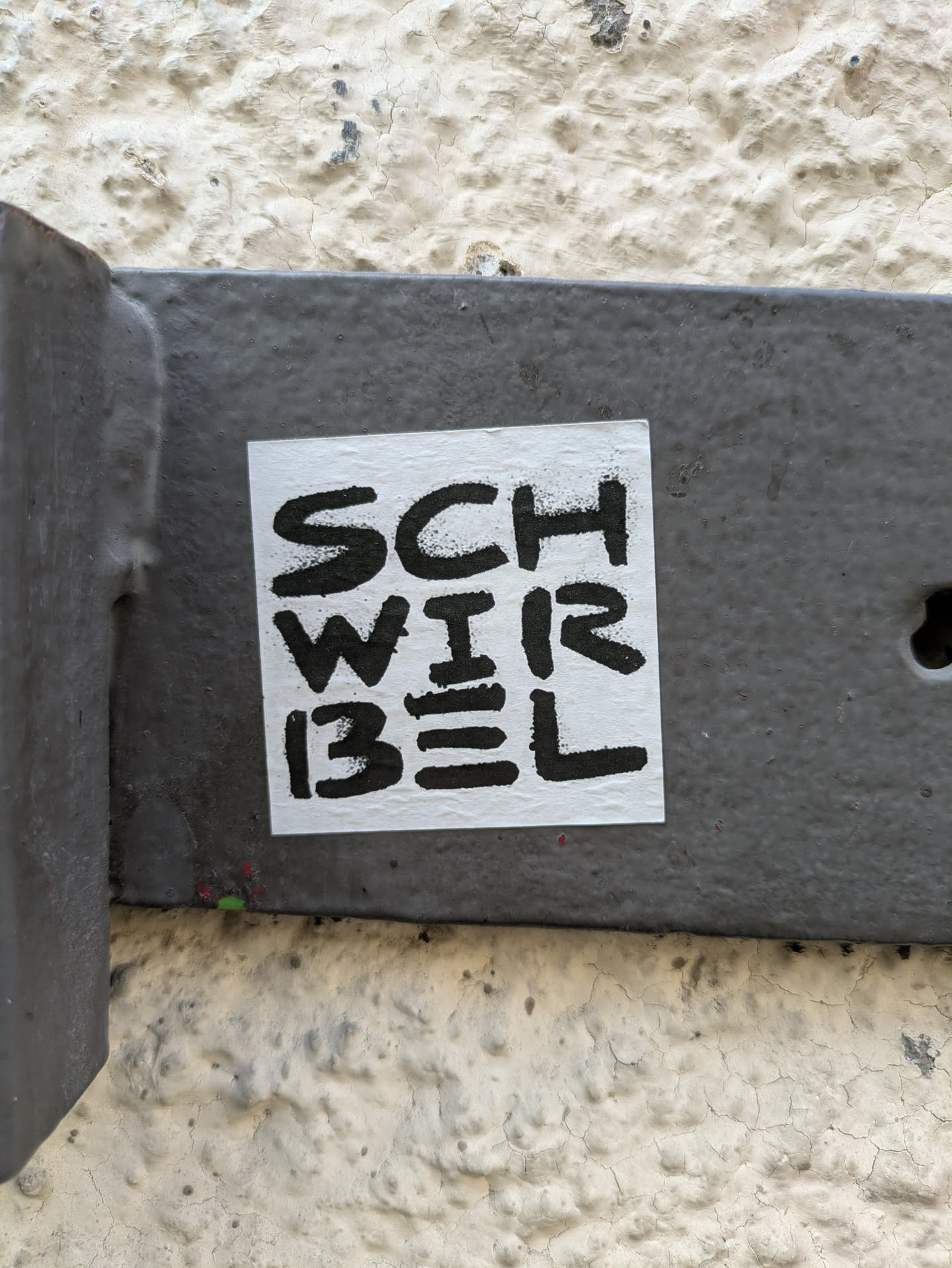 A square, white sticker with black text is adhered to a gray metal fixture. The sticker reads 'SCHWIRBEL' in a stacked format. The text is bold and slightly distressed in appearance. The sticker is centrally located in the frame, with the metal fixture providing a contrasting backdrop. The fixture is attached to a textured, light-colored wall.
