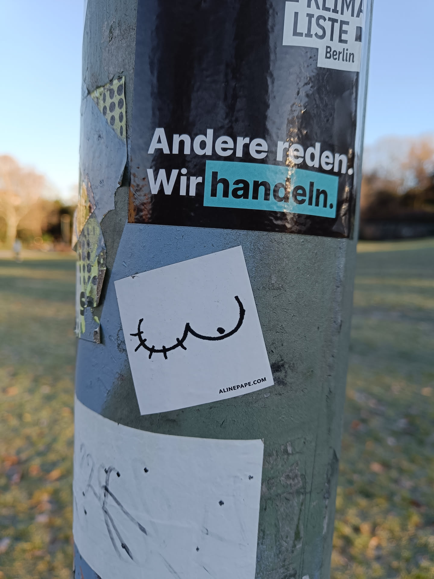 A small, minimalist sticker depicting a stylized breast with eyelashes. The sticker is affixed to a lamppost alongside other stickers, one of which reads "Andere reden. Wir handeln." (Others talk. We act.) in German.  The sticker has a website address at the bottom.