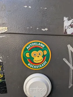 Street sticker A circular sticker with a thick yellow border. Inside is a dark green band containing the bold white text 'AUTOKLANG' curved along the top and 'ROCKSTEADY' curved along the bottom. The center features a stylized cartoon monkey head with green fur, yellow facial skin and inner ears, large wide-open eyes, and a slight smirk, outlined by a red circle.