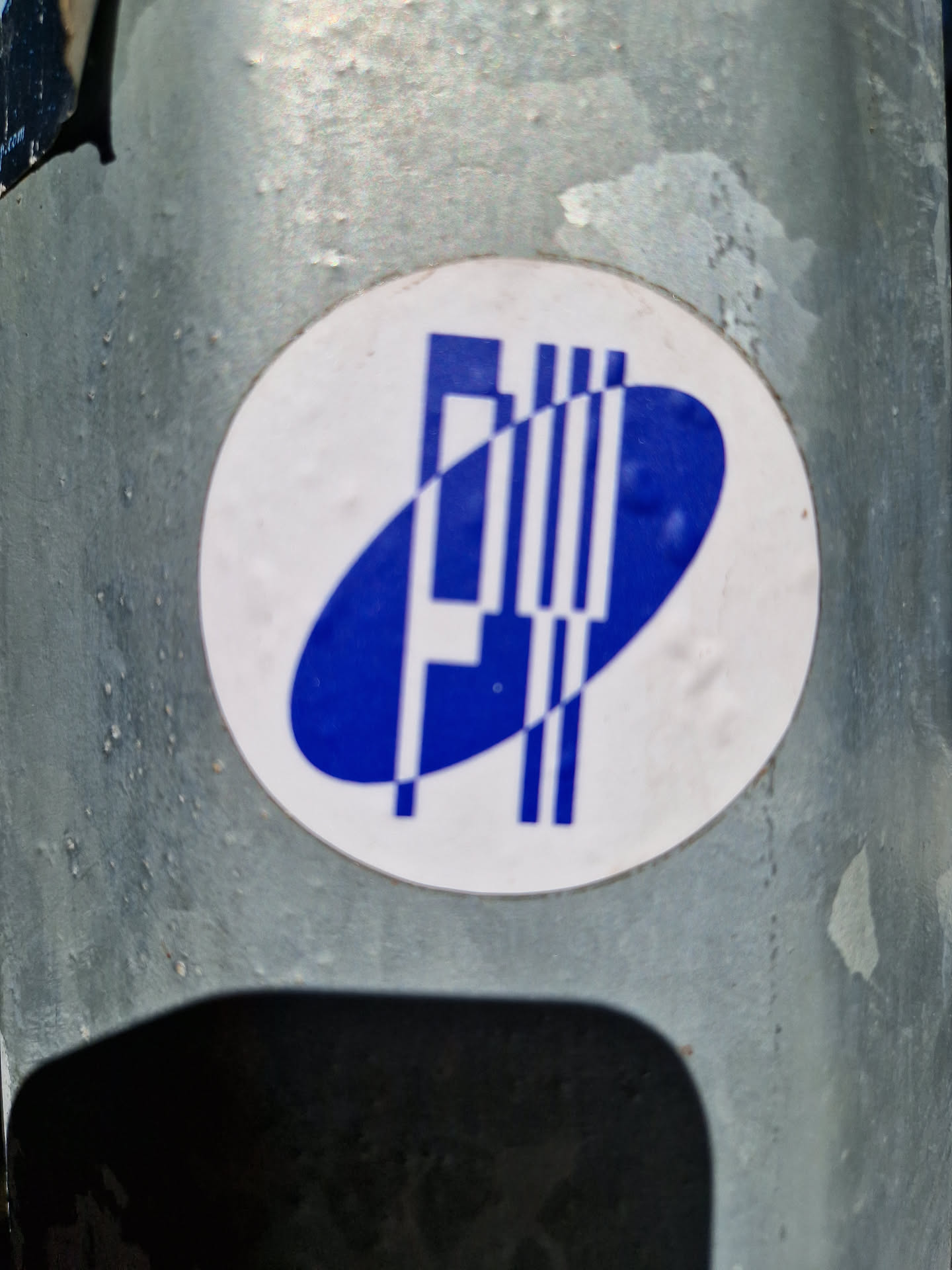 A round sticker with a blue and white logo. The logo features a stylized letter 'P' or similar form within an oval shape, accompanied by parallel lines.  The sticker appears to be affixed to a metallic surface.