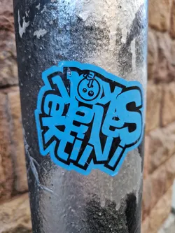 A blue and black sticker with stylized text that says "Noys Crew."  There's a small illustration of what looks like a bowling ball with a face in the middle of the design. The sticker is affixed to a dark-colored pole.