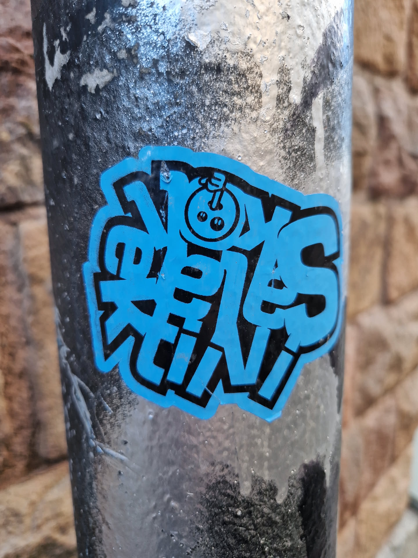 A blue and black sticker with stylized text that says "Noys Crew."  There's a small illustration of what looks like a bowling ball with a face in the middle of the design. The sticker is affixed to a dark-colored pole.