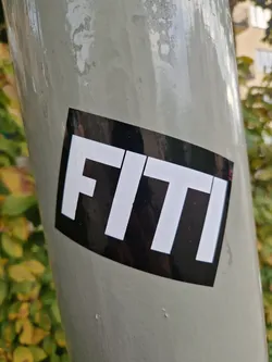 A black and white sticker with the word FITI in a bold, sans-serif font. The sticker is diamond-shaped with rounded corners and has a glossy finish. It's adhered to a light grey pole.