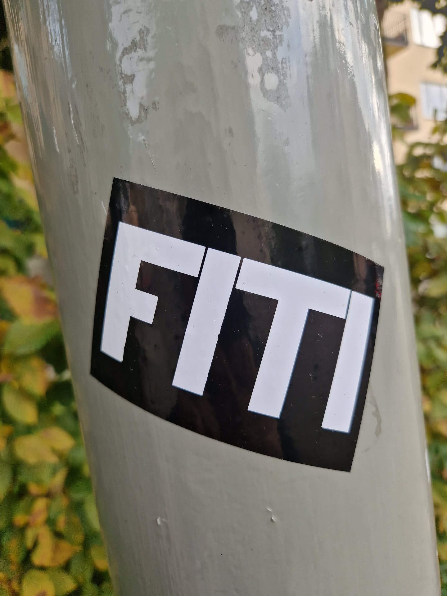 A black and white sticker with the word FITI in a bold, sans-serif font. The sticker is diamond-shaped with rounded corners and has a glossy finish. It's adhered to a light grey pole.