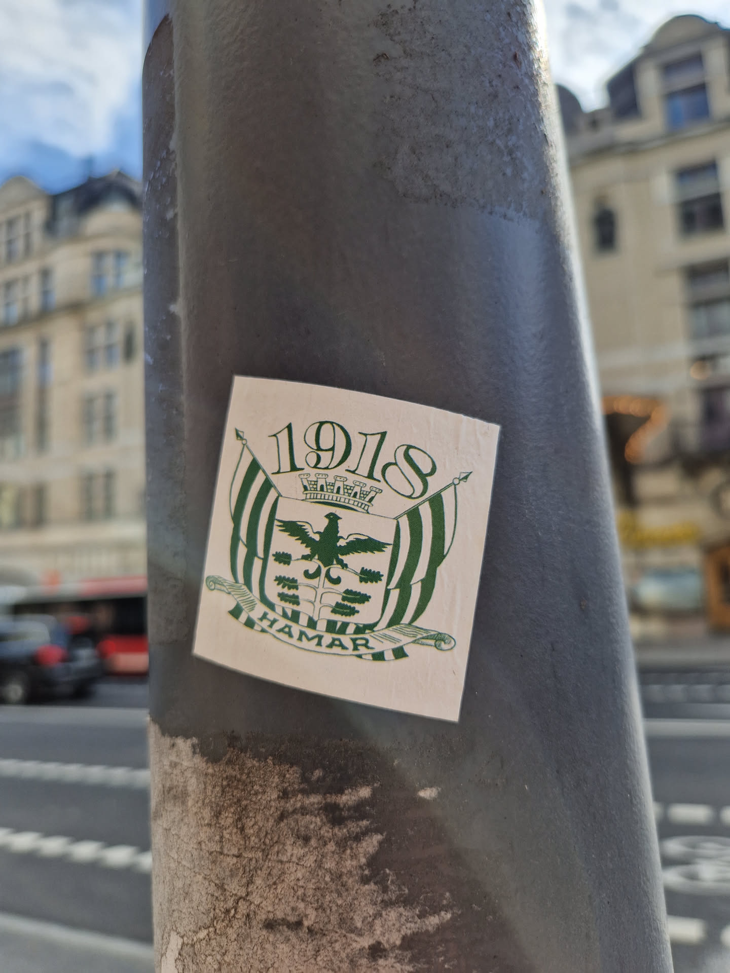 A sticker featuring the coat of arms of Hamar, Norway, with the year 1918 prominently displayed. The design is simple, using a cream background with green and black detailing. The sticker is affixed to a grey pole in an urban setting.