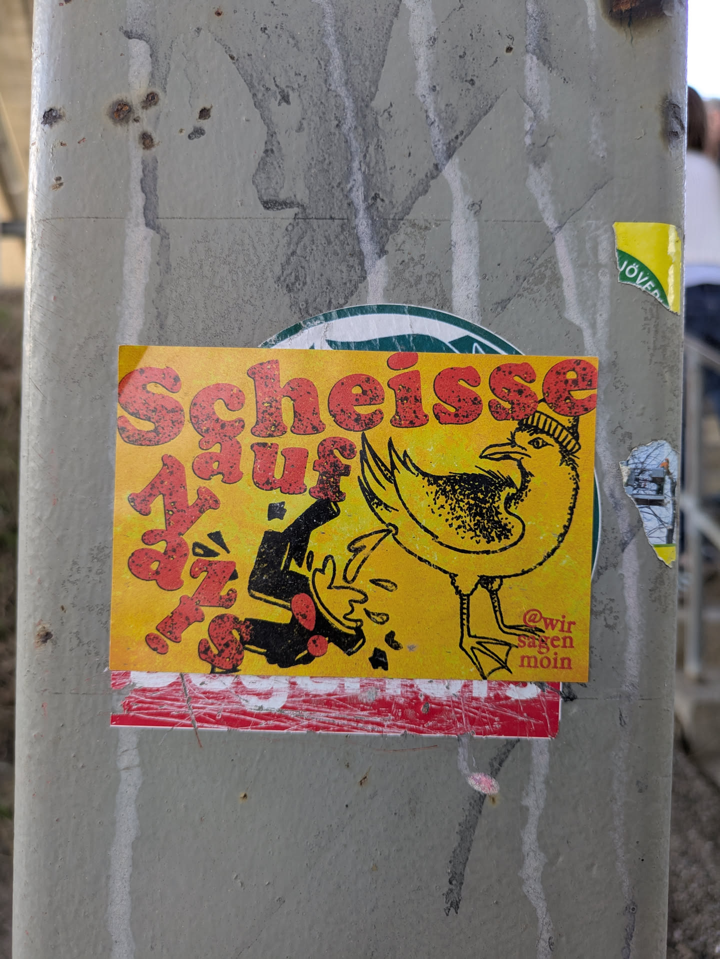 This sticker features a yellow background with red text that says "Scheisse auf Nazis." There is an image of a seagull wearing a knit cap kicking a black figure representing a Nazi, with money and tomatoes being thrown from the kicked figure. The sticker is applied on a gray concrete pole.