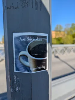 The sticker shows a photo of a cup of coffee. Text above the image reads "Arsta Skivakademi". Text below the image reads "Ha en bra dag," which translates to "Have a good day." The sticker is affixed to a grey pole outdoors.